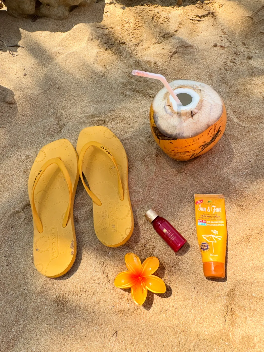 Yellow flip-flops, a king coconut, sunscreen and a frangipani flower laid out on the sand