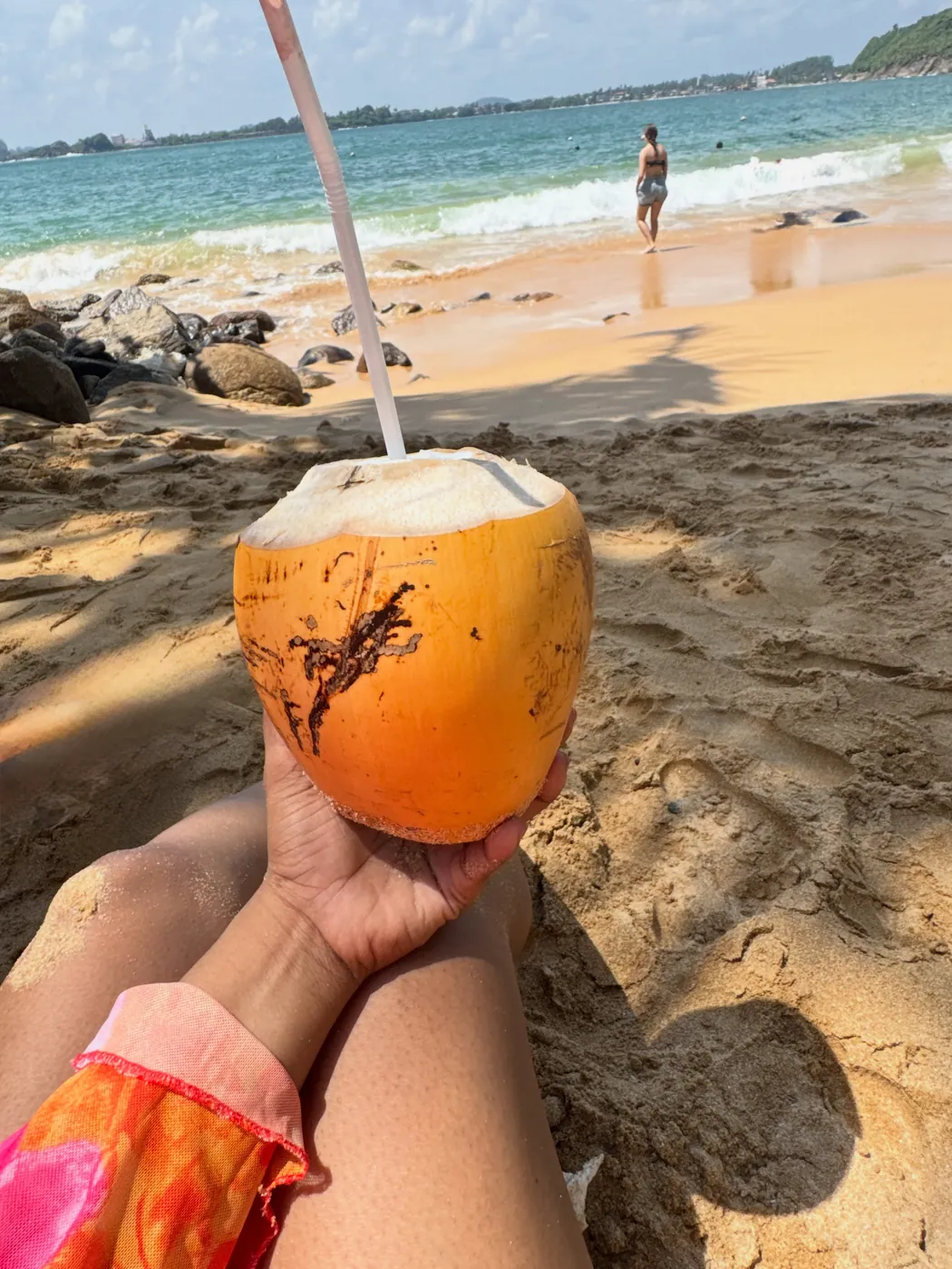 Hand holding a king coconut with a straw on the sand at Jungle Beach with the sea behind